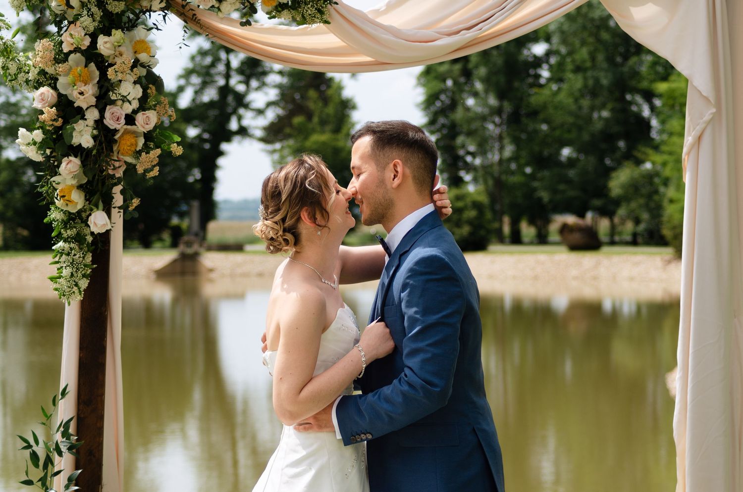 Wedding ceremony under draped arch with floral arrangements by lakeside.