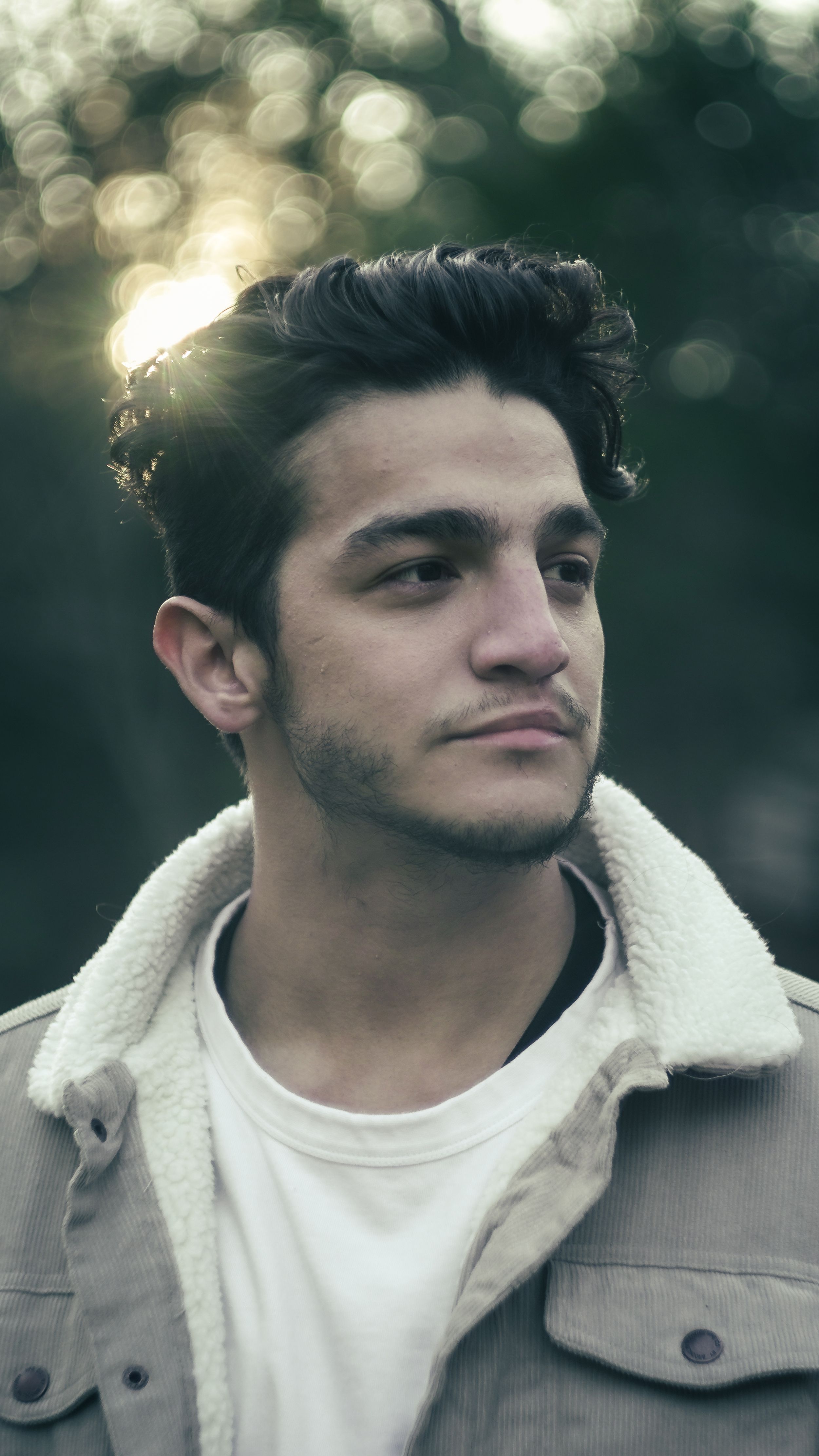 A young person in a tan jacket with sherpa lining looks to the side against a bokeh background at sunset.