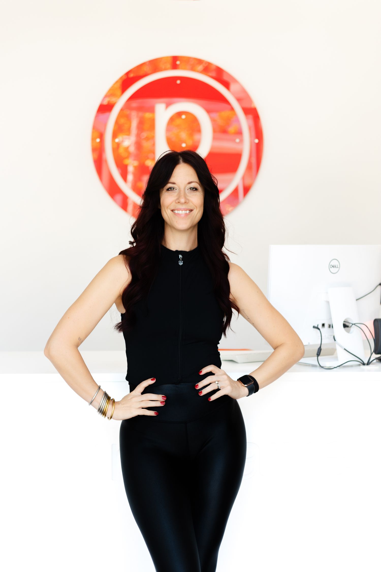 Person in black athletic wear poses confidently in front of red circular logo on white wall.