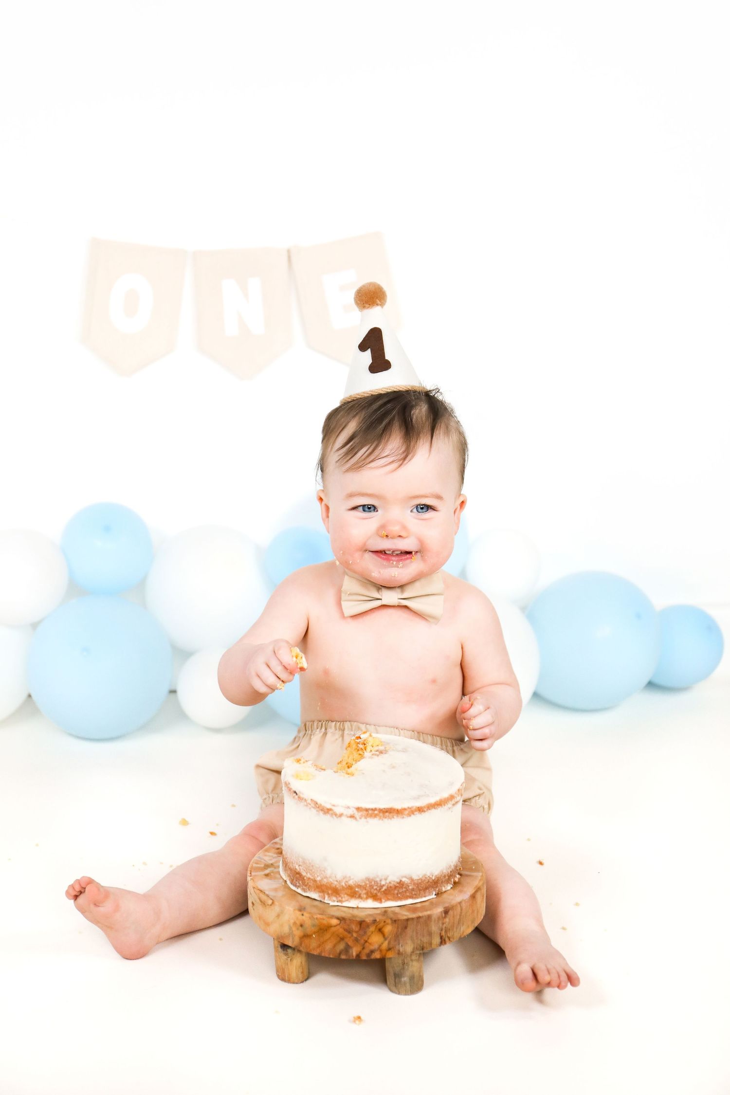 Baby in party hat sits with white cake on wooden stand surrounded by blue balloons for first birthday celebration.