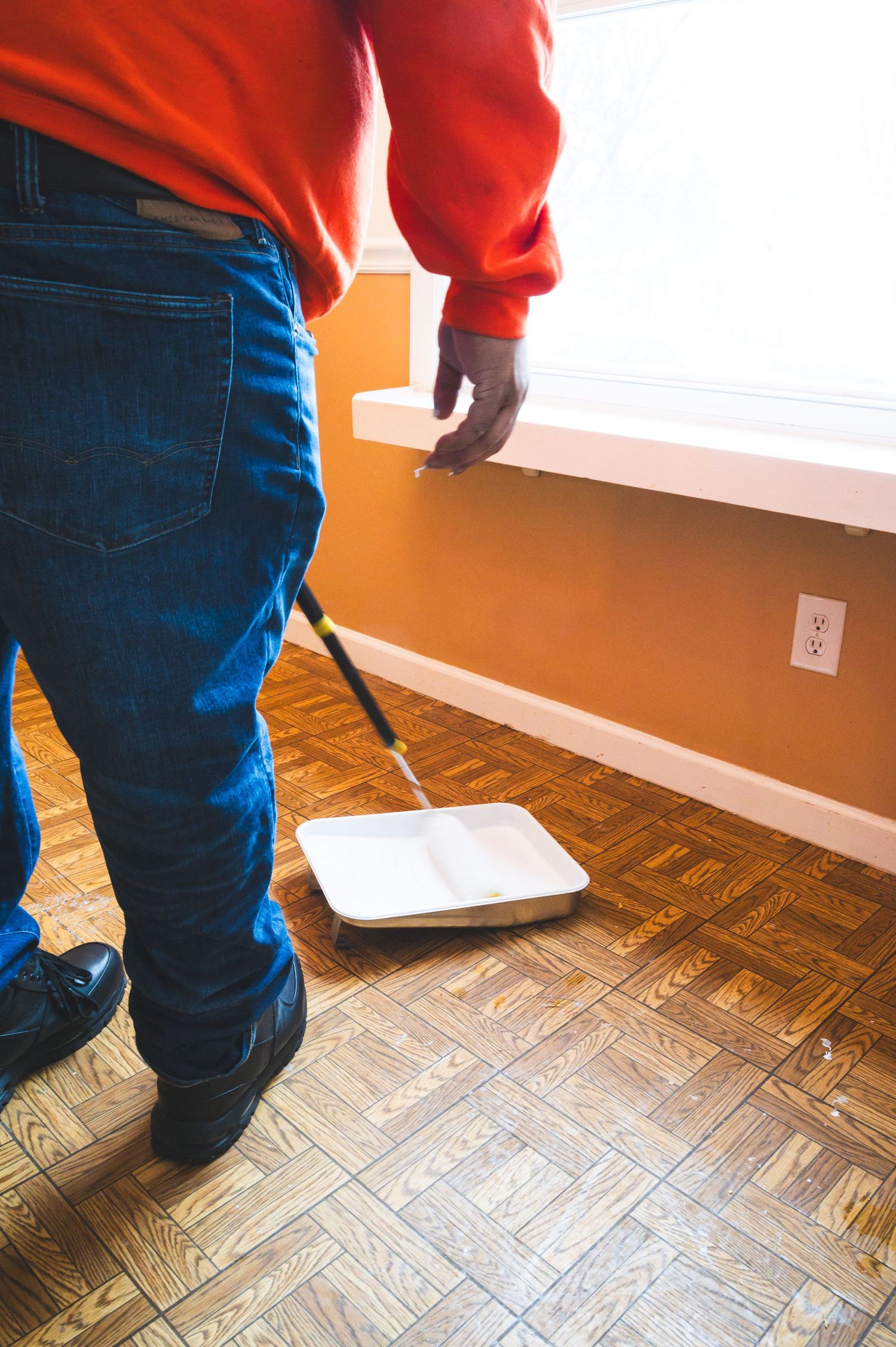 Person in orange sweater and jeans painting baseboards with a roller brush.