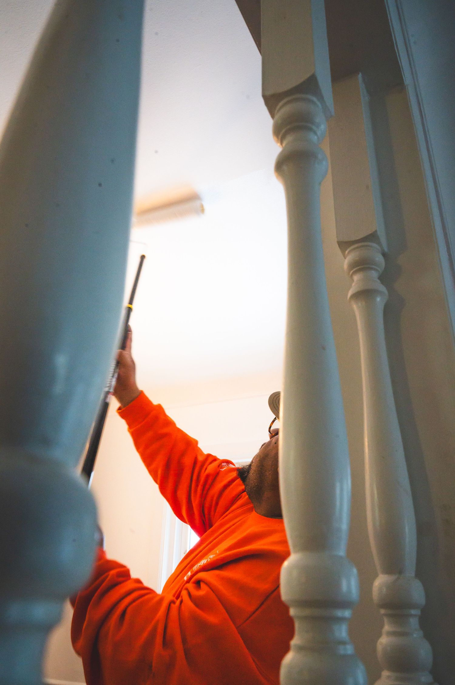 Worker in orange sweater reaching up to paint between white staircase spindles.