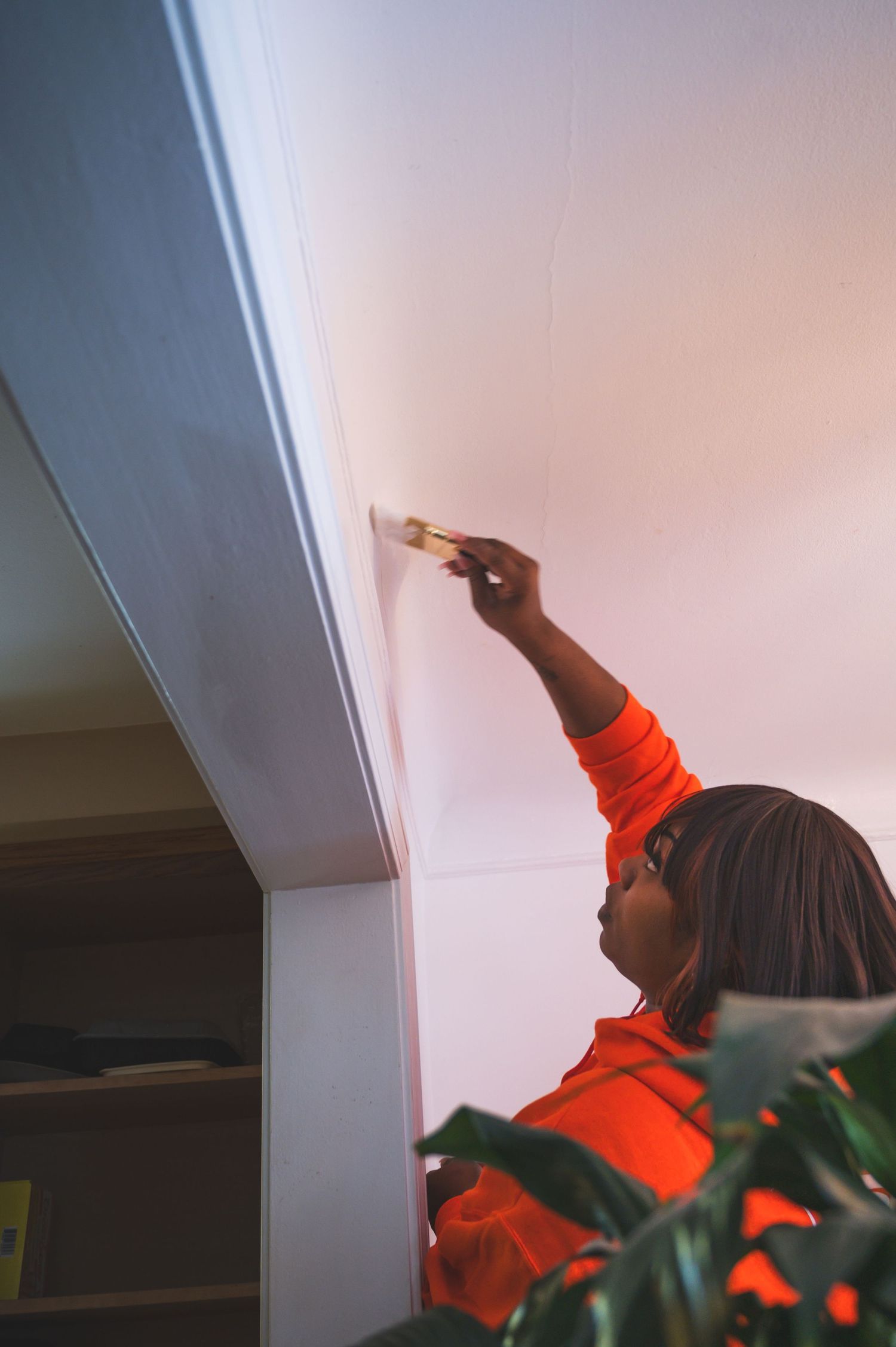 Person in orange sweater reaching up to paint a ceiling corner with careful precision.