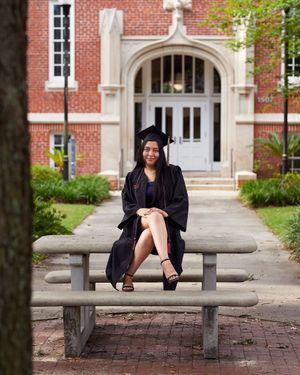 Graduate in cap and gown sits on concrete bench in front of brick university building.