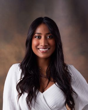 Professional headshot against soft brown backdrop featuring warm smile and flowing dark hair.