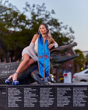 Graduate posing with decorative alligator statue while wearing a blue graduation stole and casual attire.
