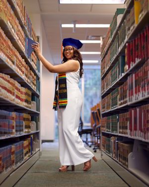 Graduate in academic regalia and white dress poses elegantly in library stacks.
