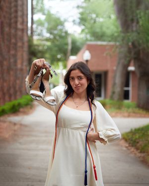 Graduate in white dress holds graduation heels on University of Florida campus walkway.