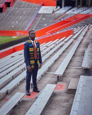 Graduate in dark suit with cultural stoles stands in stadium bleachers.