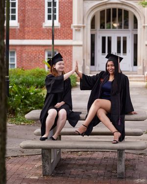 Two graduates in black caps and gowns high-five while sitting on campus bench.