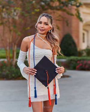Graduate holding mortarboard in white dress with red and blue honor cords.