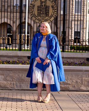 Graduate in blue academic regalia poses in front of university seal and iron gates.