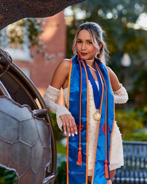 Graduate in white dress with blue and orange honor cords posing near decorative sculpture.