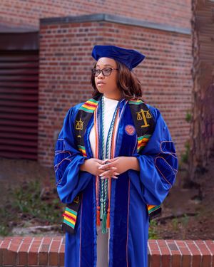 Graduate in blue doctoral regalia with colorful stoles stands proudly against brick wall.