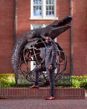 Stylishly dressed person posing with large dinosaur sculpture against brick building backdrop.