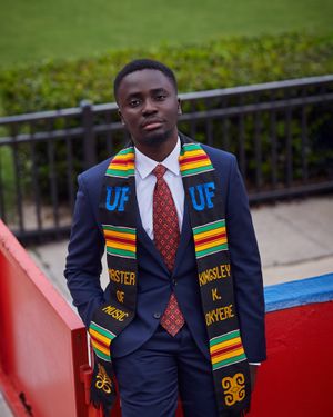 Graduate wearing navy suit and colorful stole poses by railing outdoors.