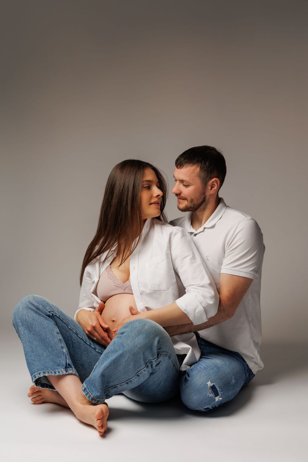 Intimate maternity portrait of couple sitting together on floor in casual wear.