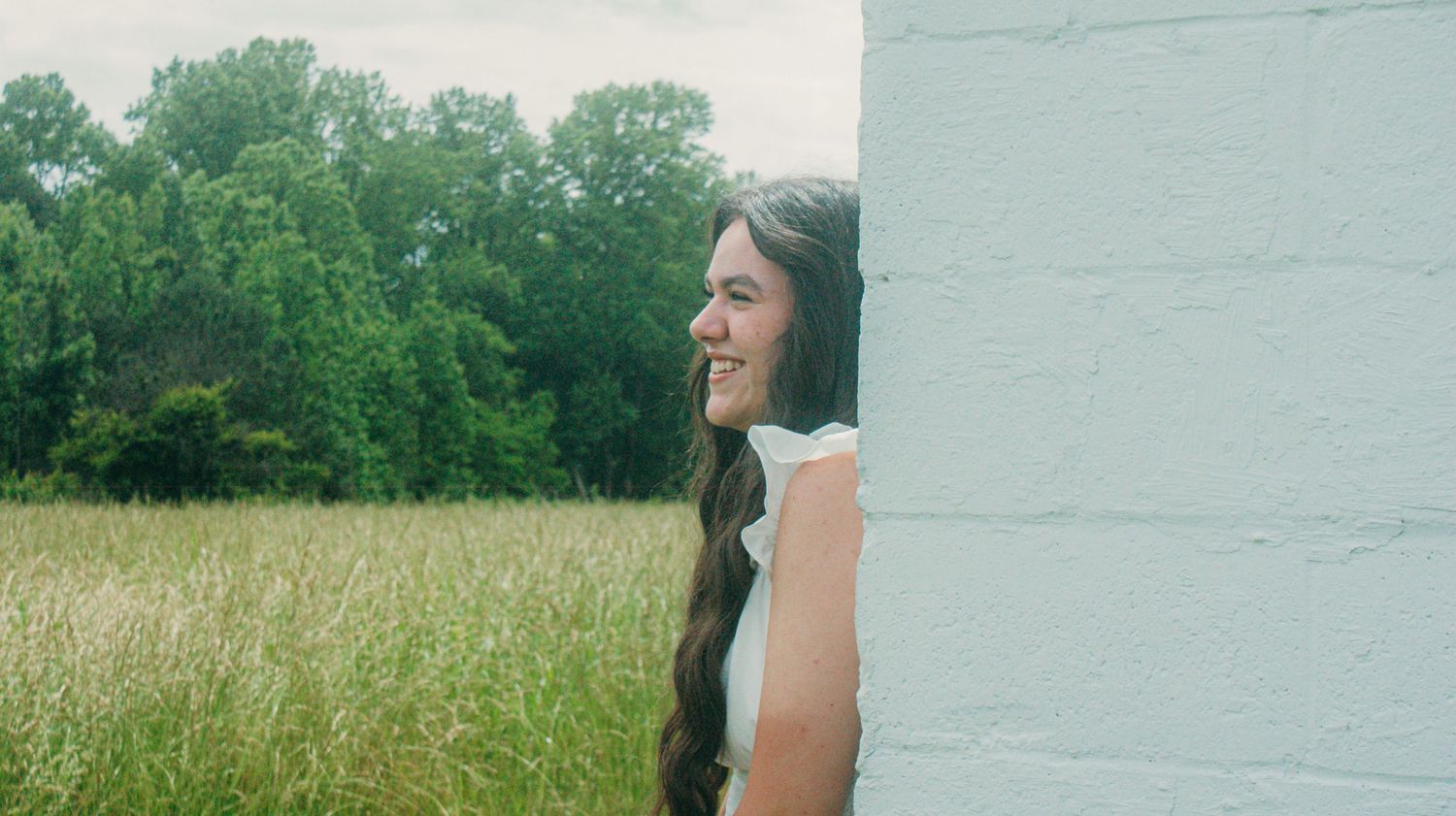 A woman in a white top stands next to a white wall overlooking a green field on a sunny day.