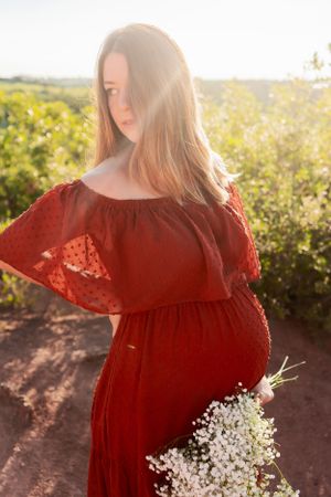A red maternity dress catches golden sunlight while holding baby's breath flowers in a natural setting.