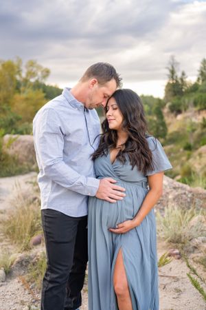 Maternity photo of couple in blue dress and white shirt standing on rocky terrain at sunset.