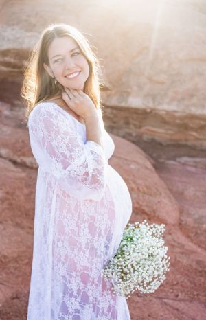A white lace maternity gown glows in golden sunlight while holding delicate baby's breath flowers.