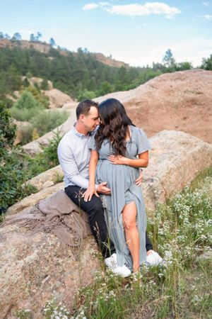 Expectant couple share intimate moment on rocky hillside wearing coordinated blue and white outfits.