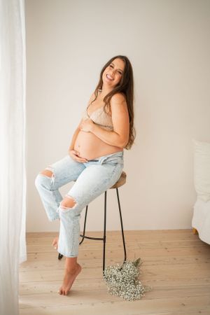 Casual maternity portrait on stool with distressed denim and neutral bralette against white wall.