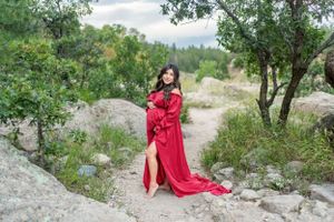 Pregnant woman in flowing red dress poses dramatically on rocky outdoor terrain.