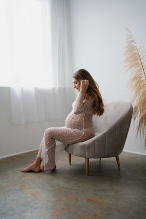 Dreamy indoor maternity portrait with woman in lace dress seated in modern gray chair.