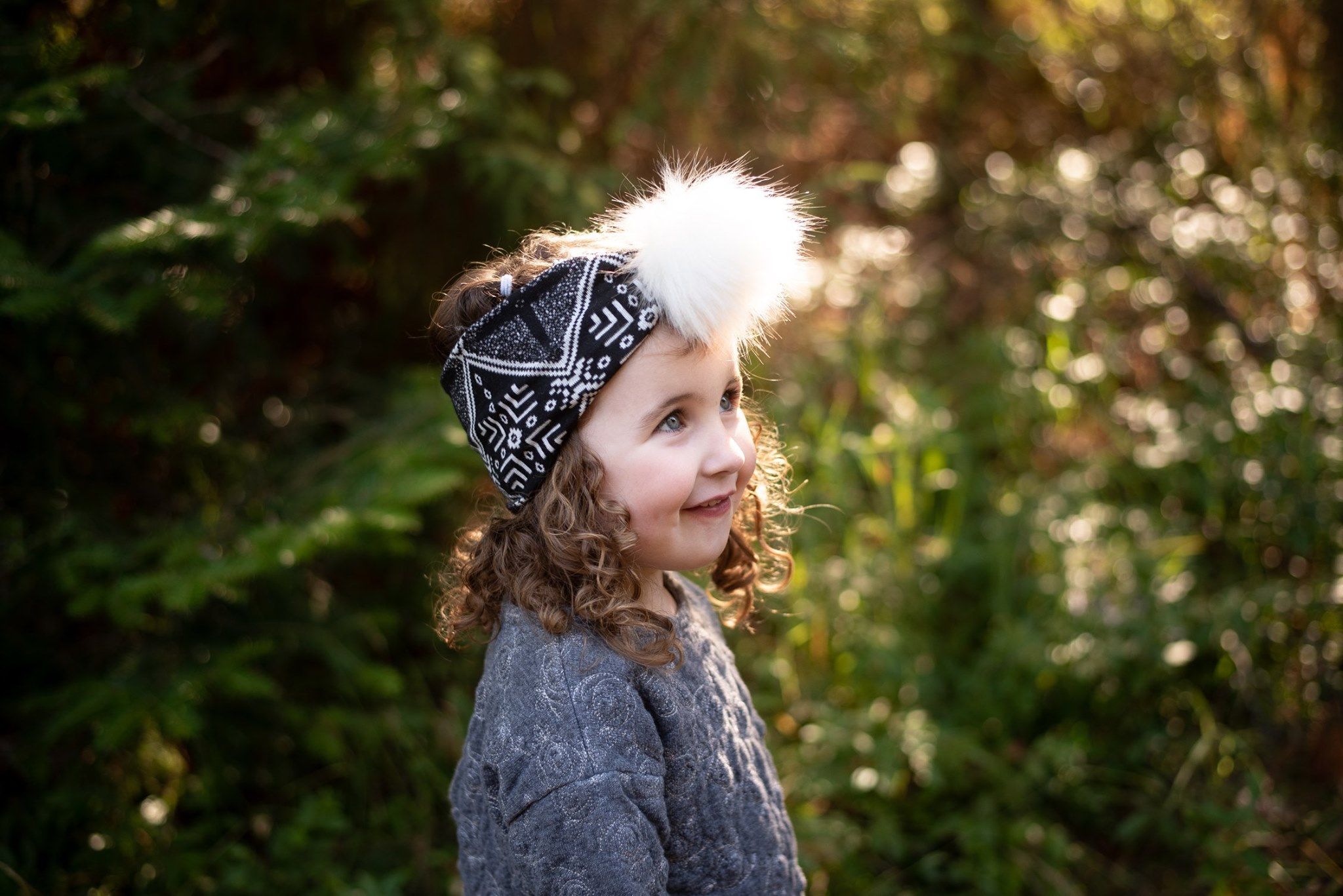 Child wearing grey sweater and bandana headband smiles in golden sunlit forest.