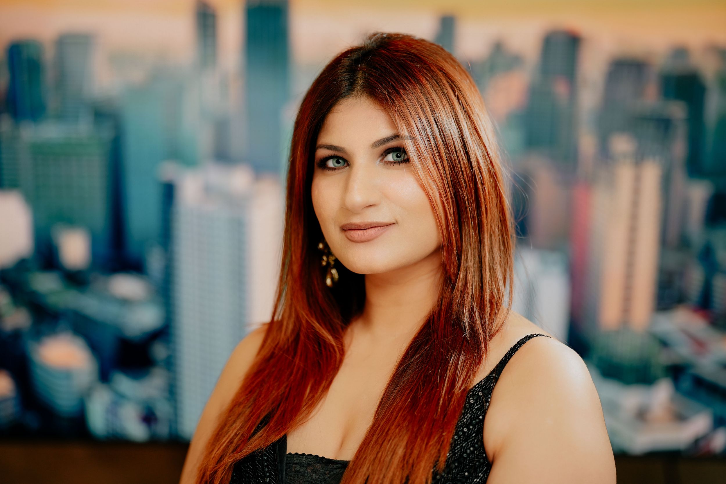 A young woman in black attire poses against a blurred cityscape background of tall buildings at sunset.