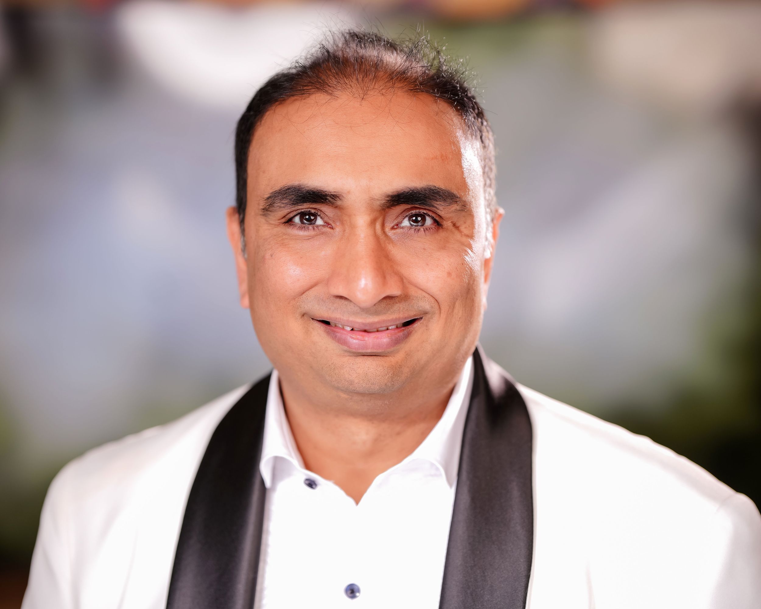 Professional headshot of someone in a white dress shirt and black shawl collar against a blurred background.