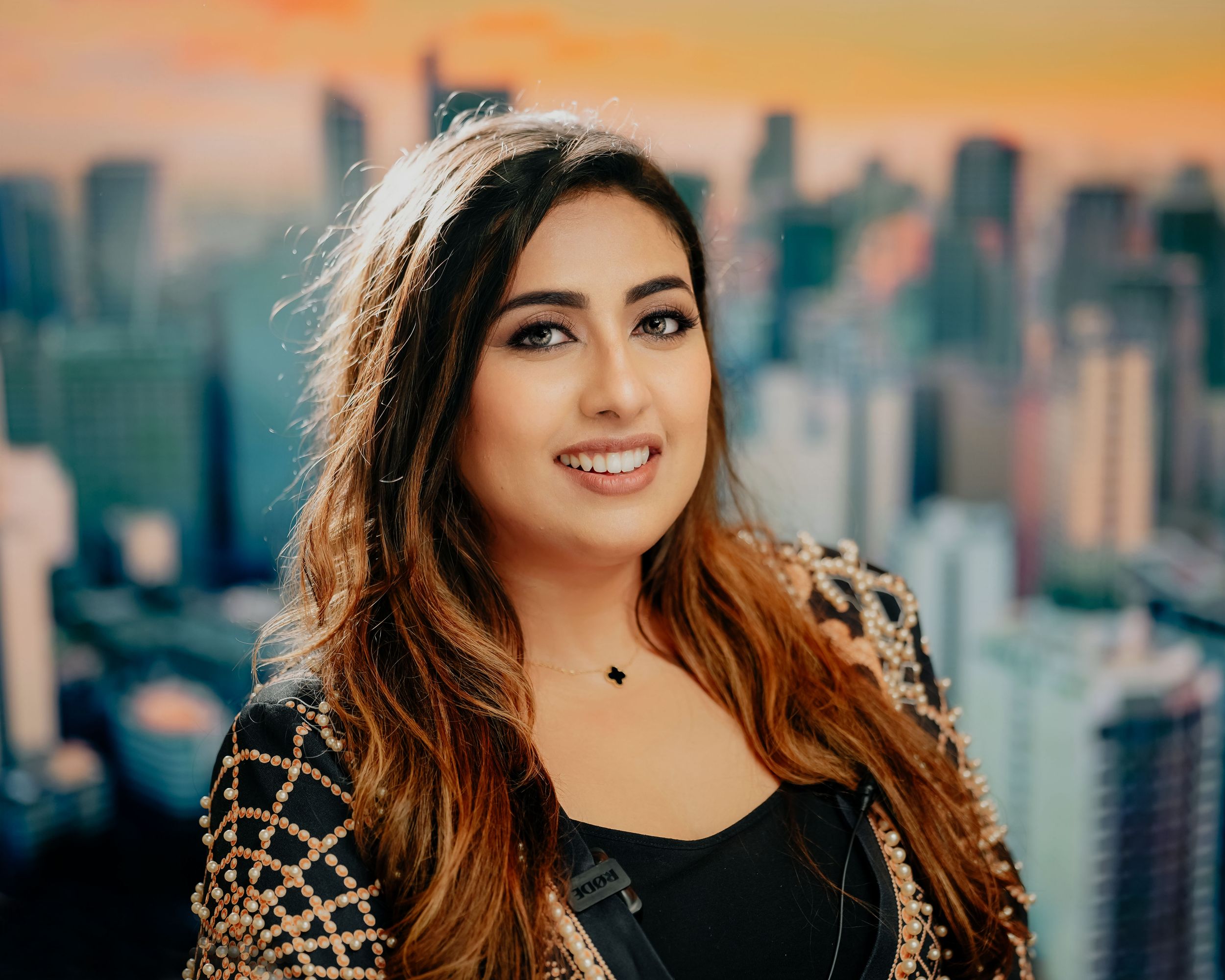 A smiling person poses against a city skyline backdrop during sunset, wearing a black top with ombre hair.