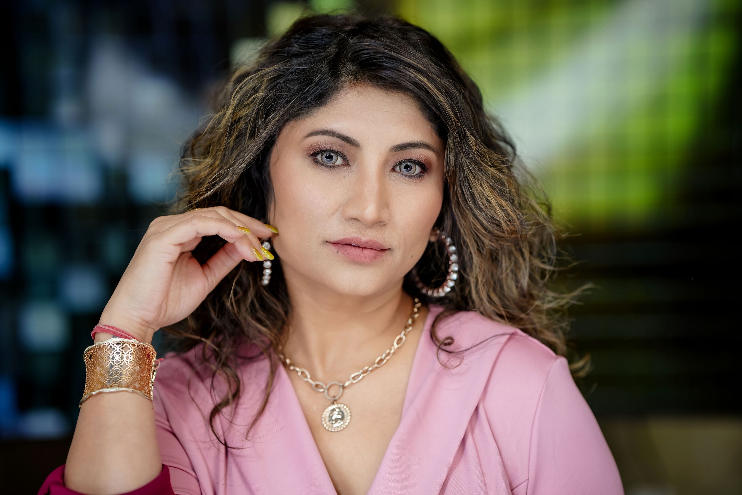 Woman wearing pink blazer and gold jewelry poses with wavy hair against blurred background.