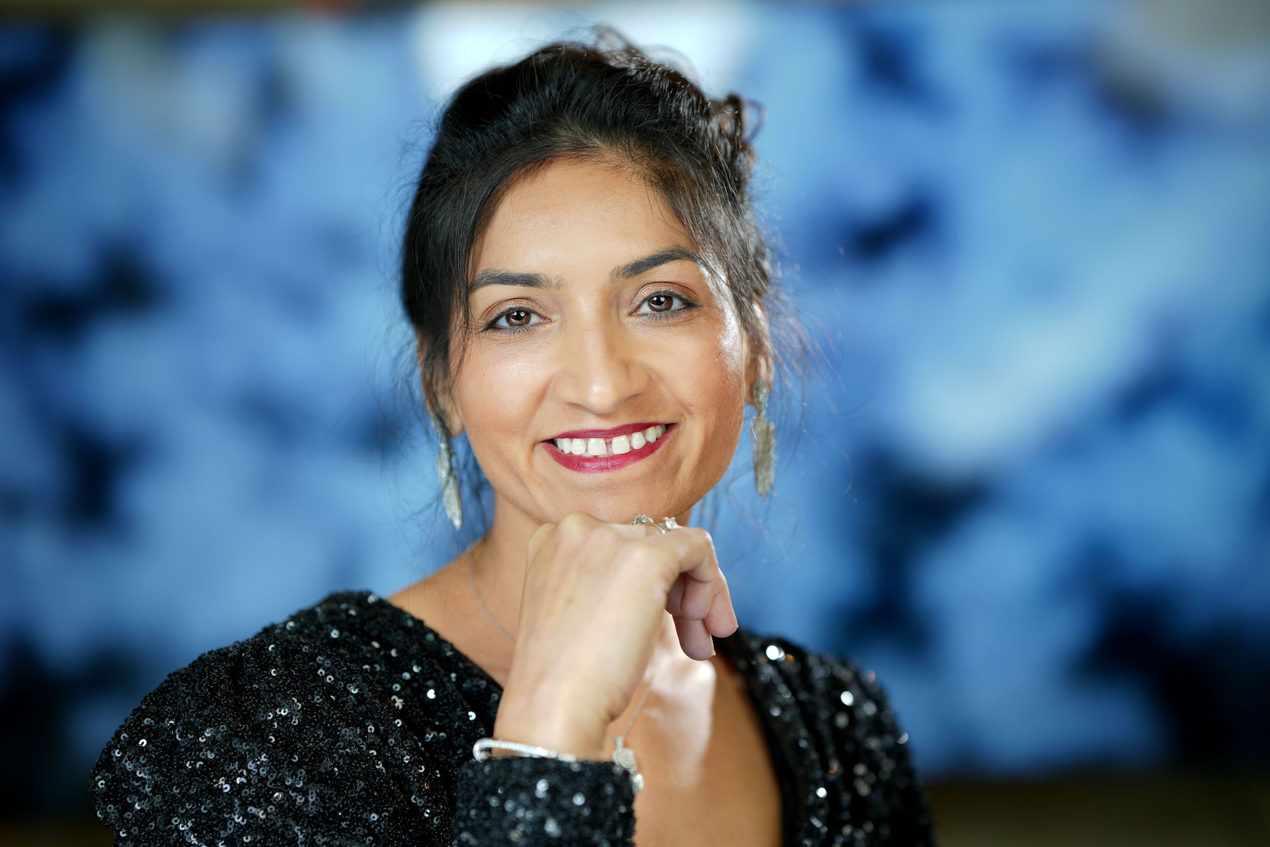 A person wearing a sparkling black top smiles warmly while resting their chin on their hand against a blue background.