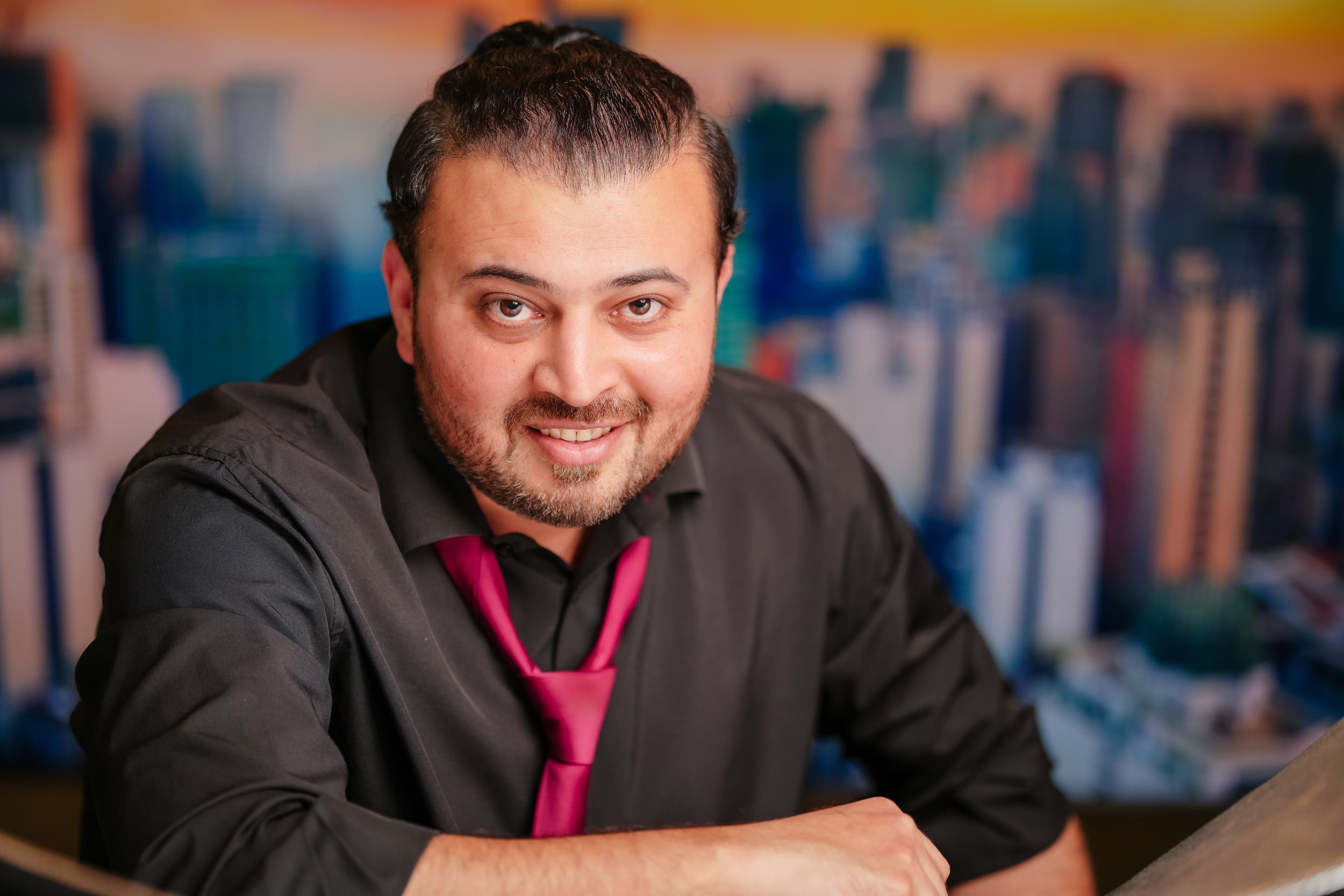 Professional headshot of someone in a black shirt and red tie against a blurred cityscape background.
