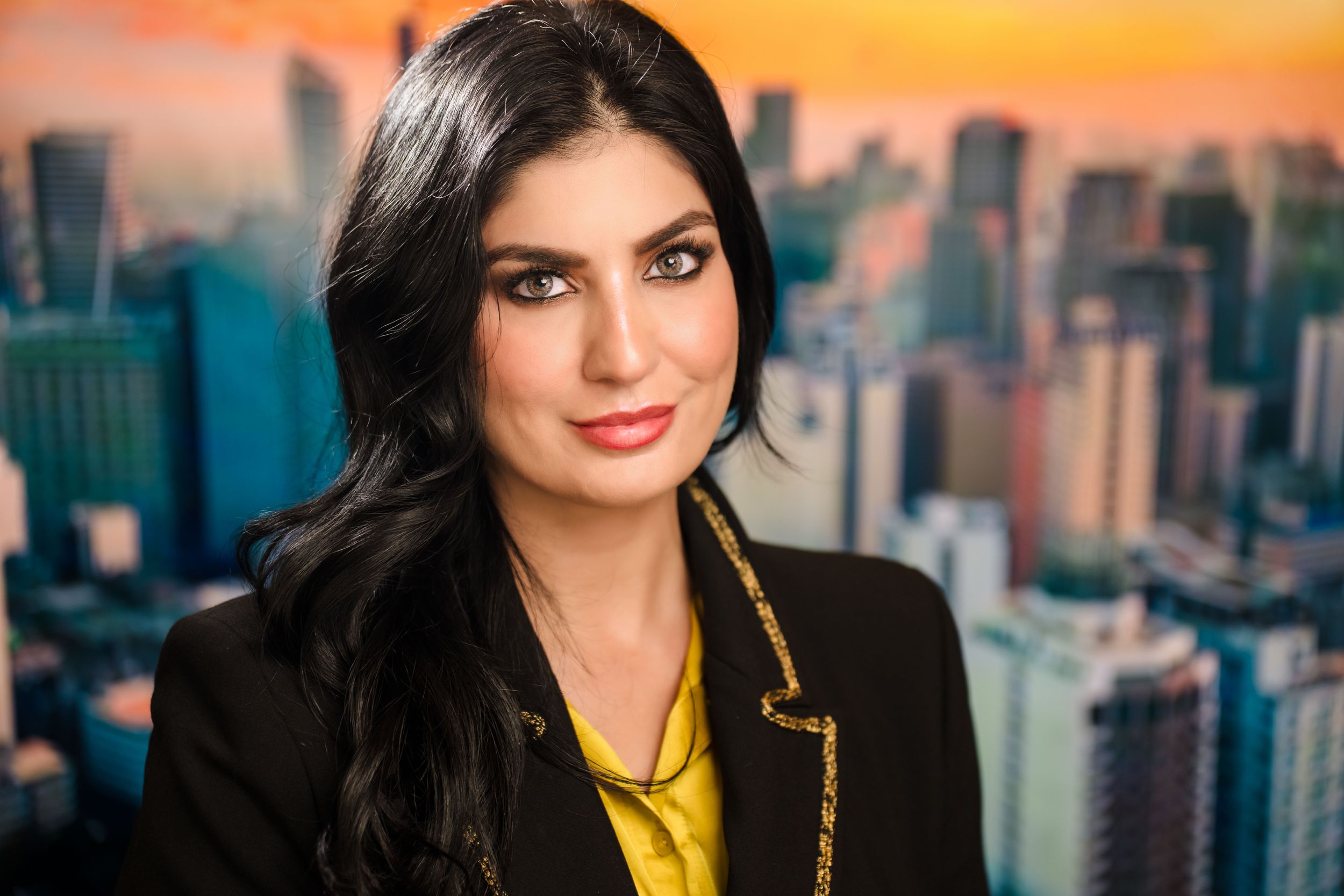Professional wearing black blazer and yellow blouse stands against backdrop of urban skyline during golden hour.