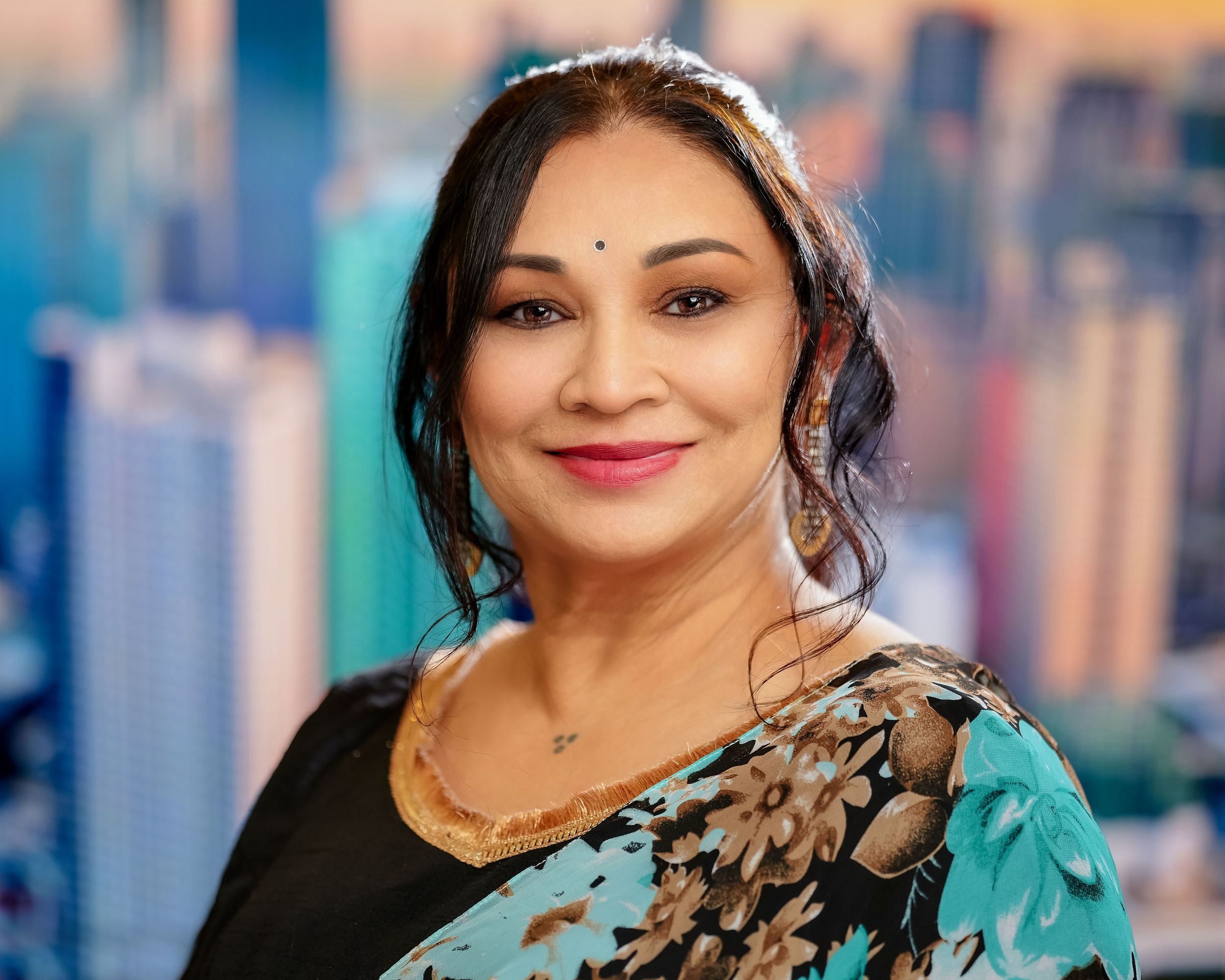 Woman in a teal and black floral dress smiles warmly against a blurred city backdrop.