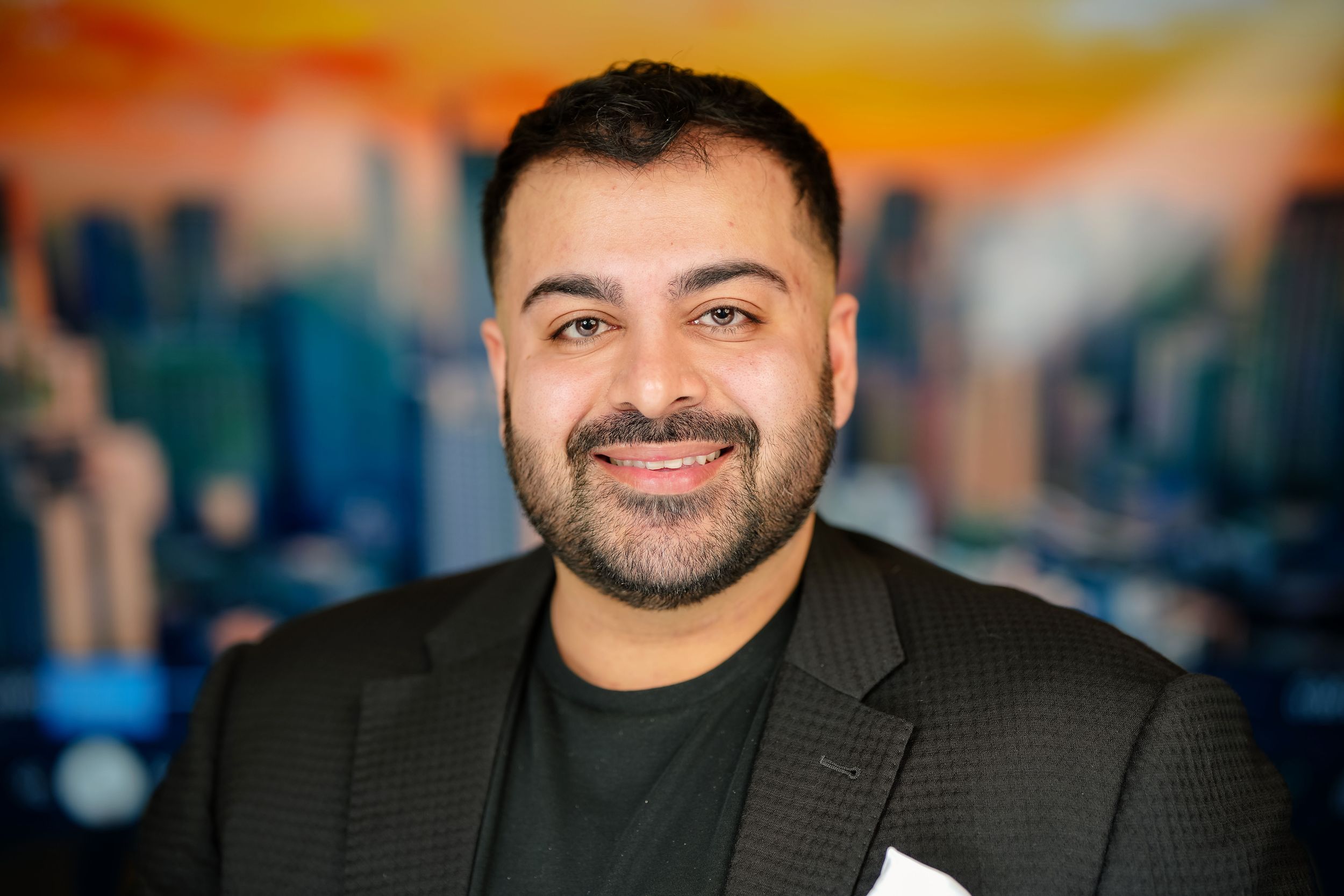 A smiling person wearing a black jacket poses against a blurred cityscape background with warm sunset lighting.