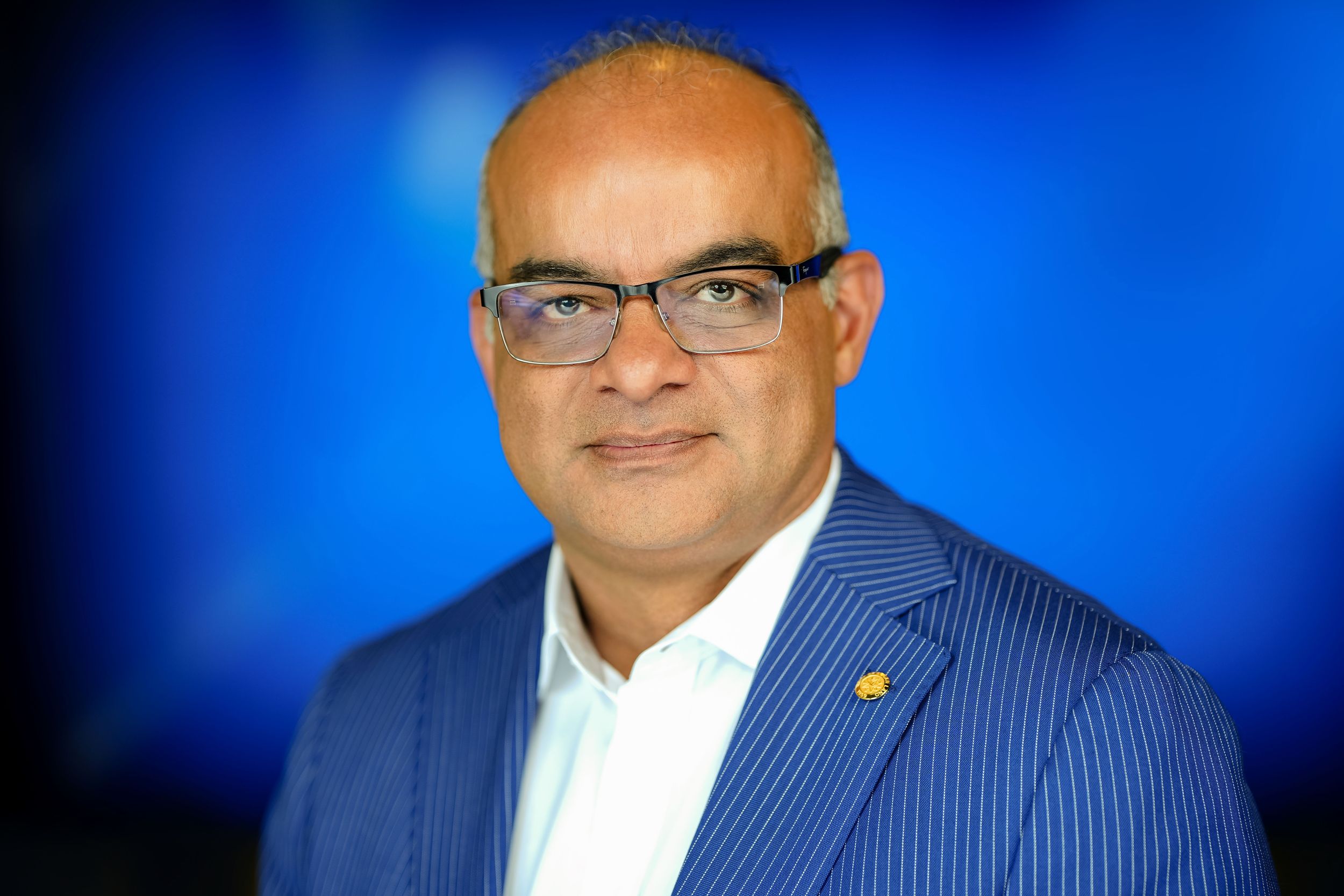 Professional headshot of person wearing glasses and blue pinstriped suit against a blurred blue background.
