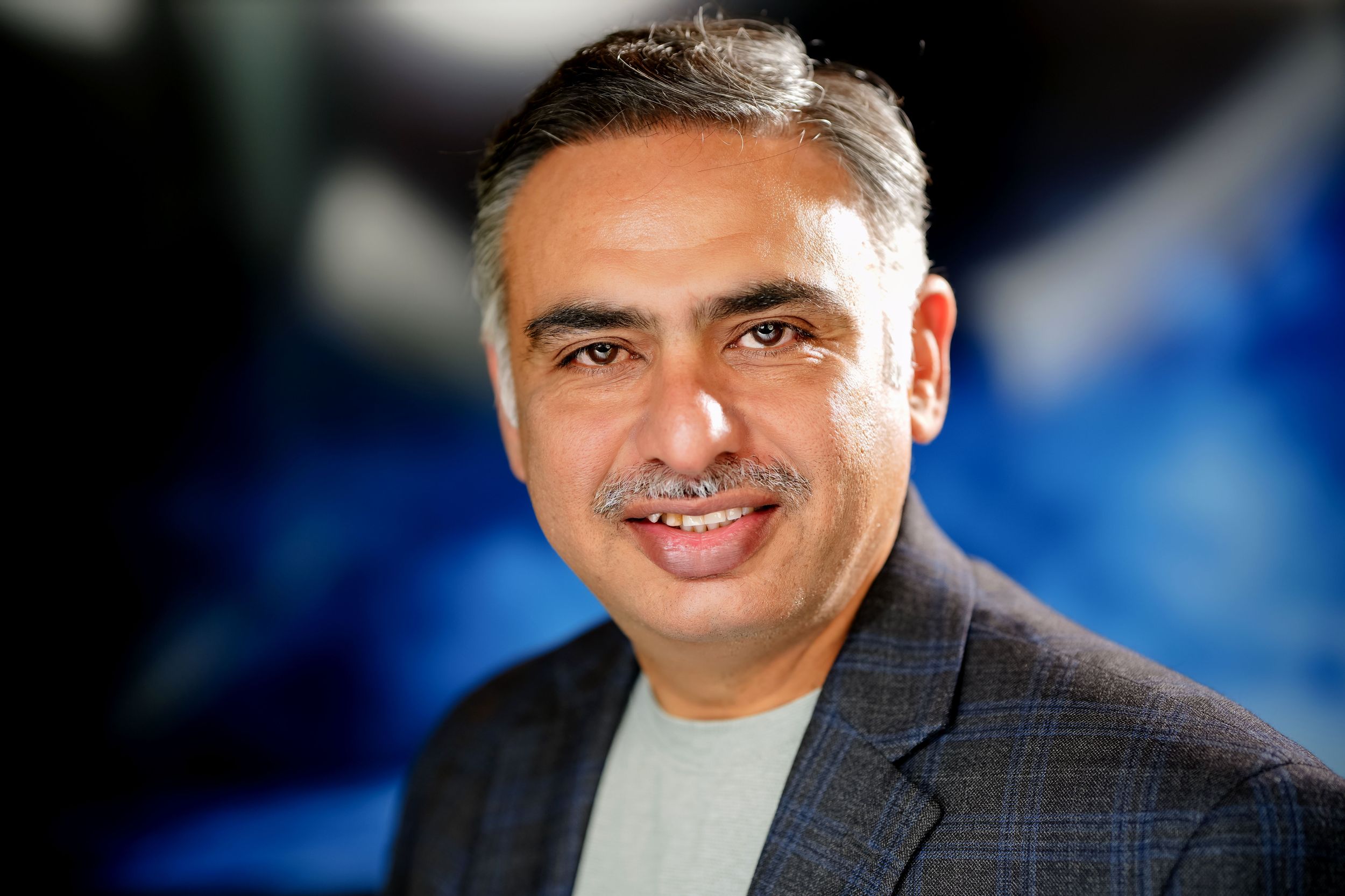 Professional headshot of someone wearing a dark suit jacket against a blue background.