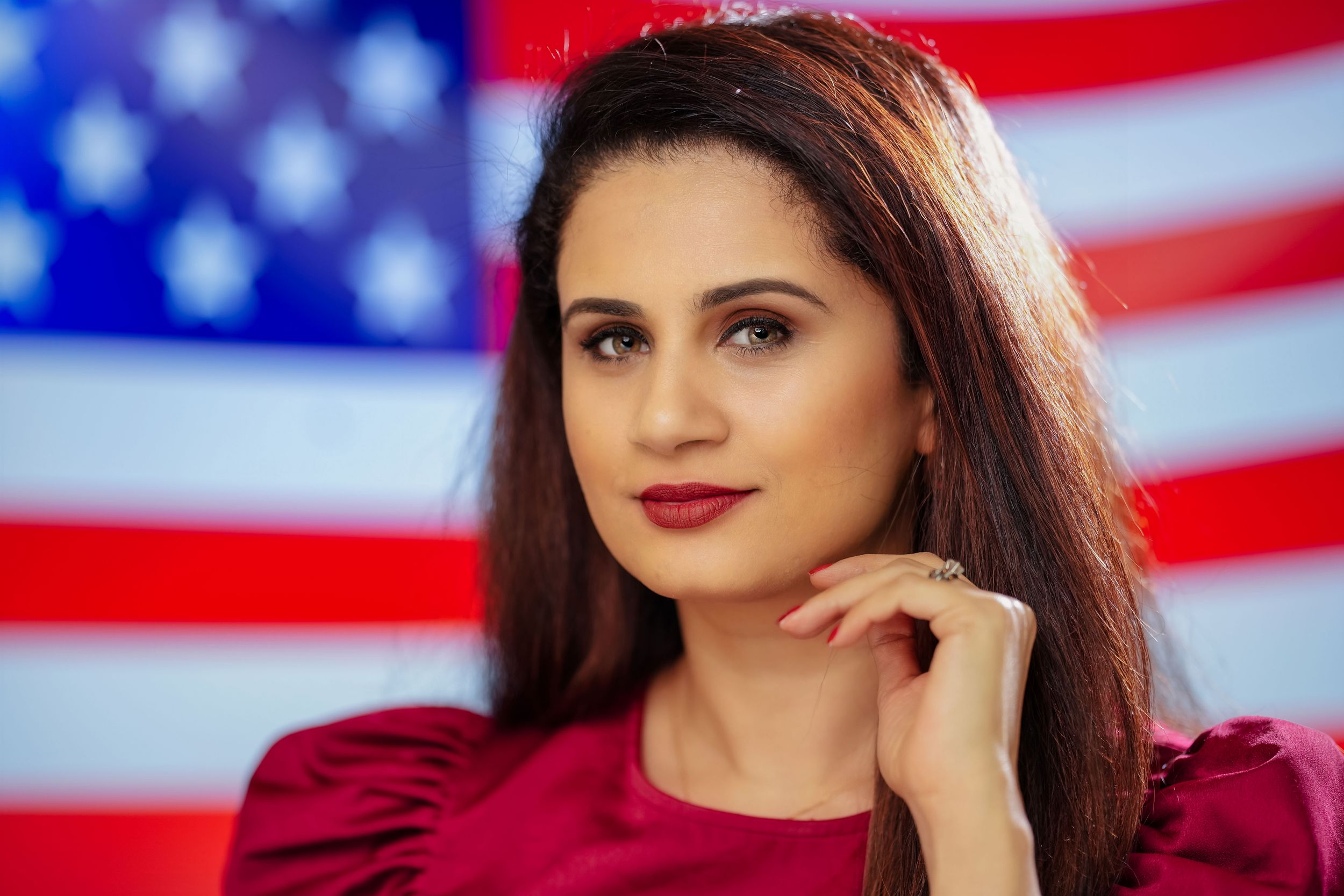 A portrait of someone in a red blouse posing in front of an American flag backdrop.