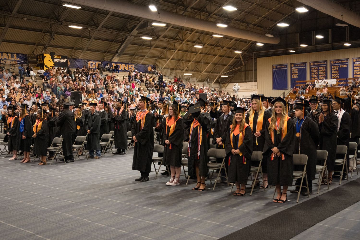 Large graduation ceremony crowd in indoor venue with graduates in caps and gowns.