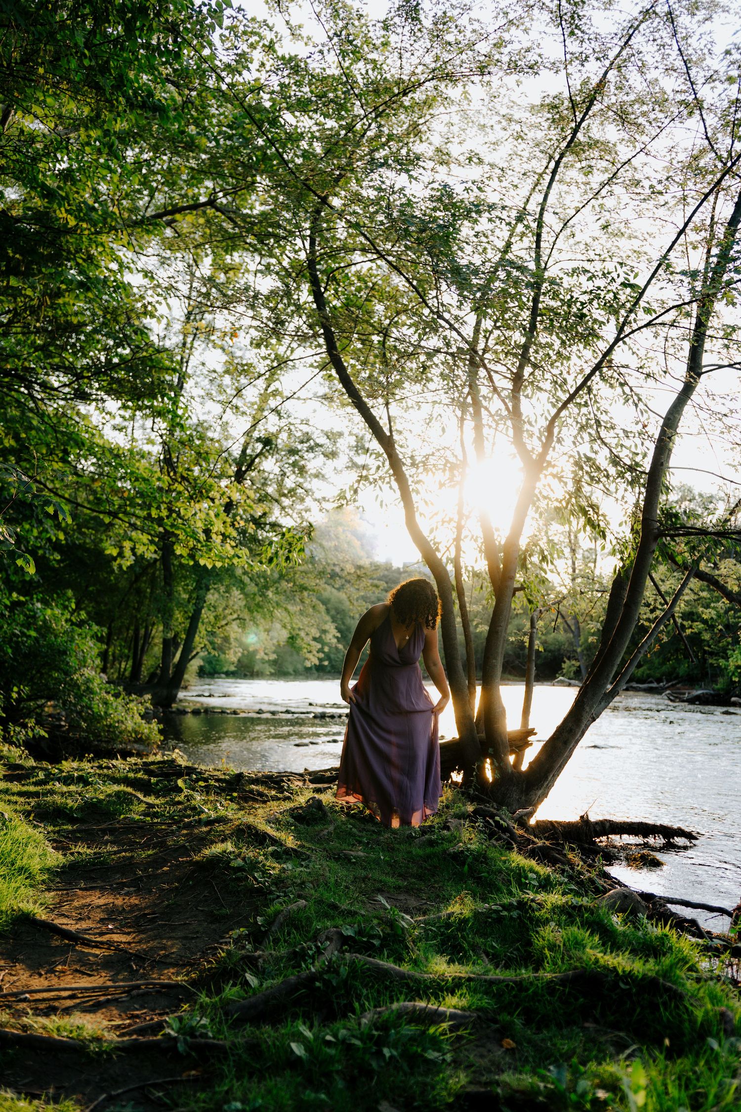 Silhouetted figure in dress by riverside at sunset with trees creating dramatic natural lighting.