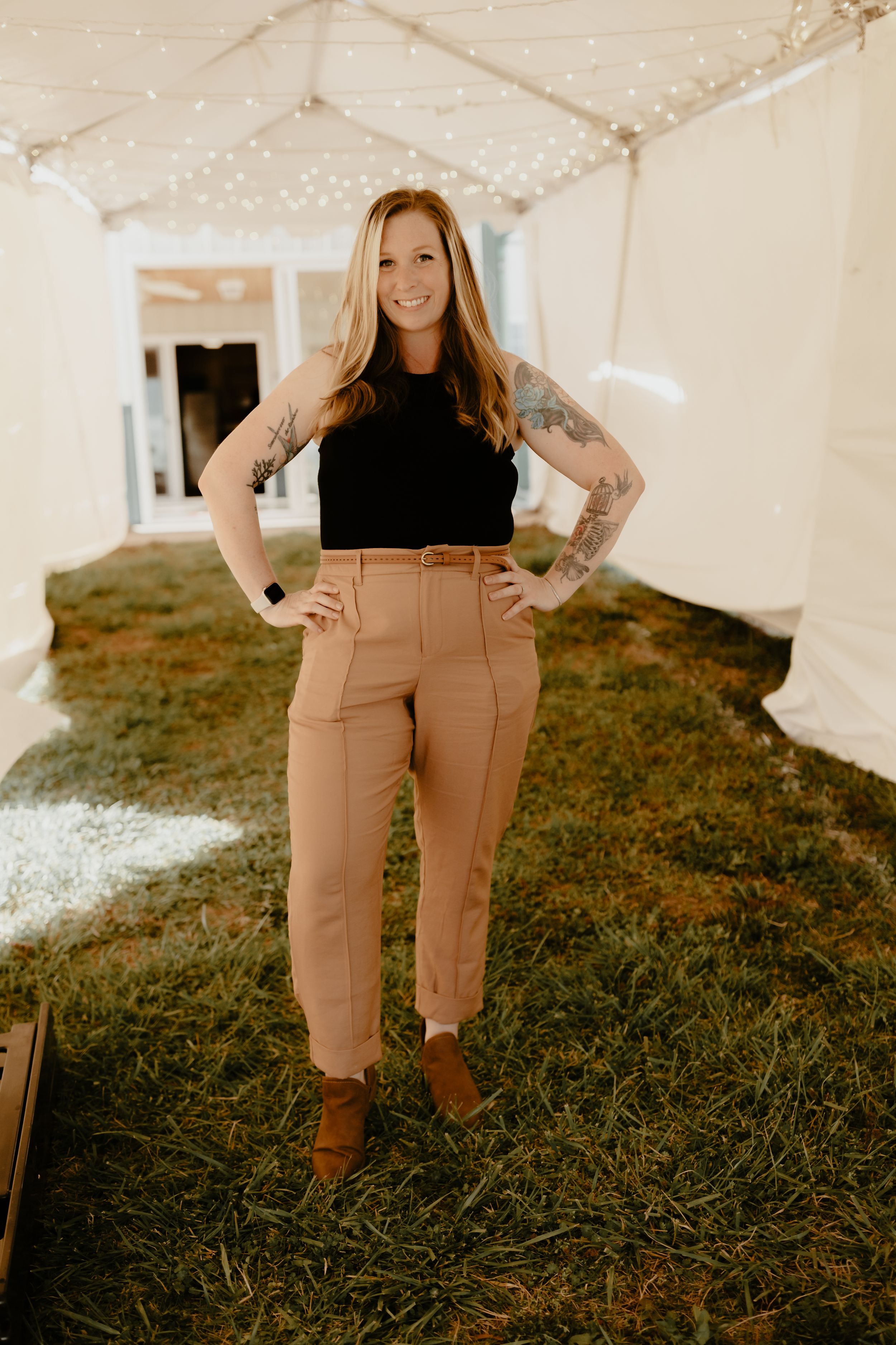 A smiling tattooed woman poses confidently in tan trousers inside a tent with string lights.