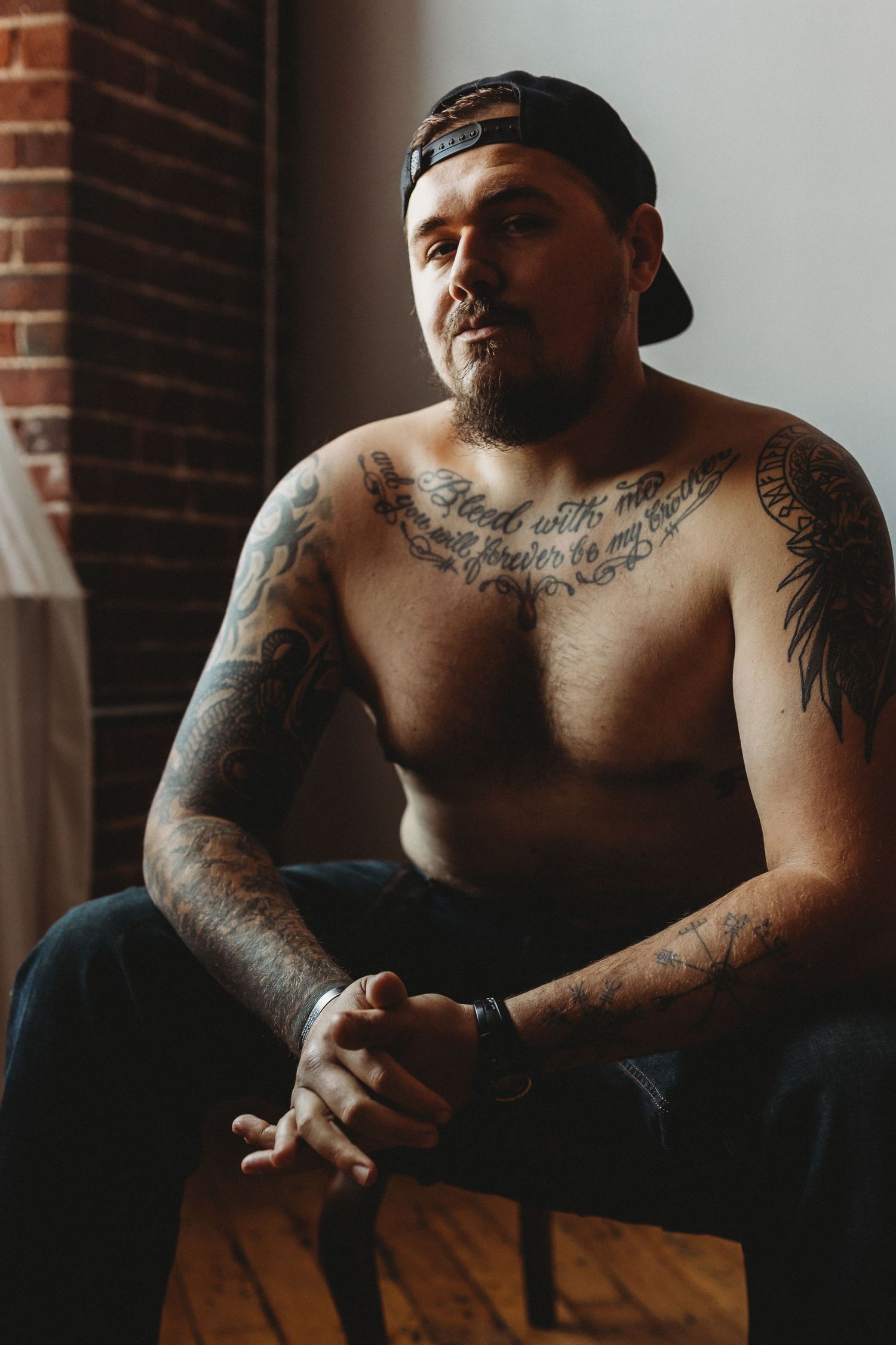 Shirtless person with intricate chest tattoos sits contemplatively by brick wall.