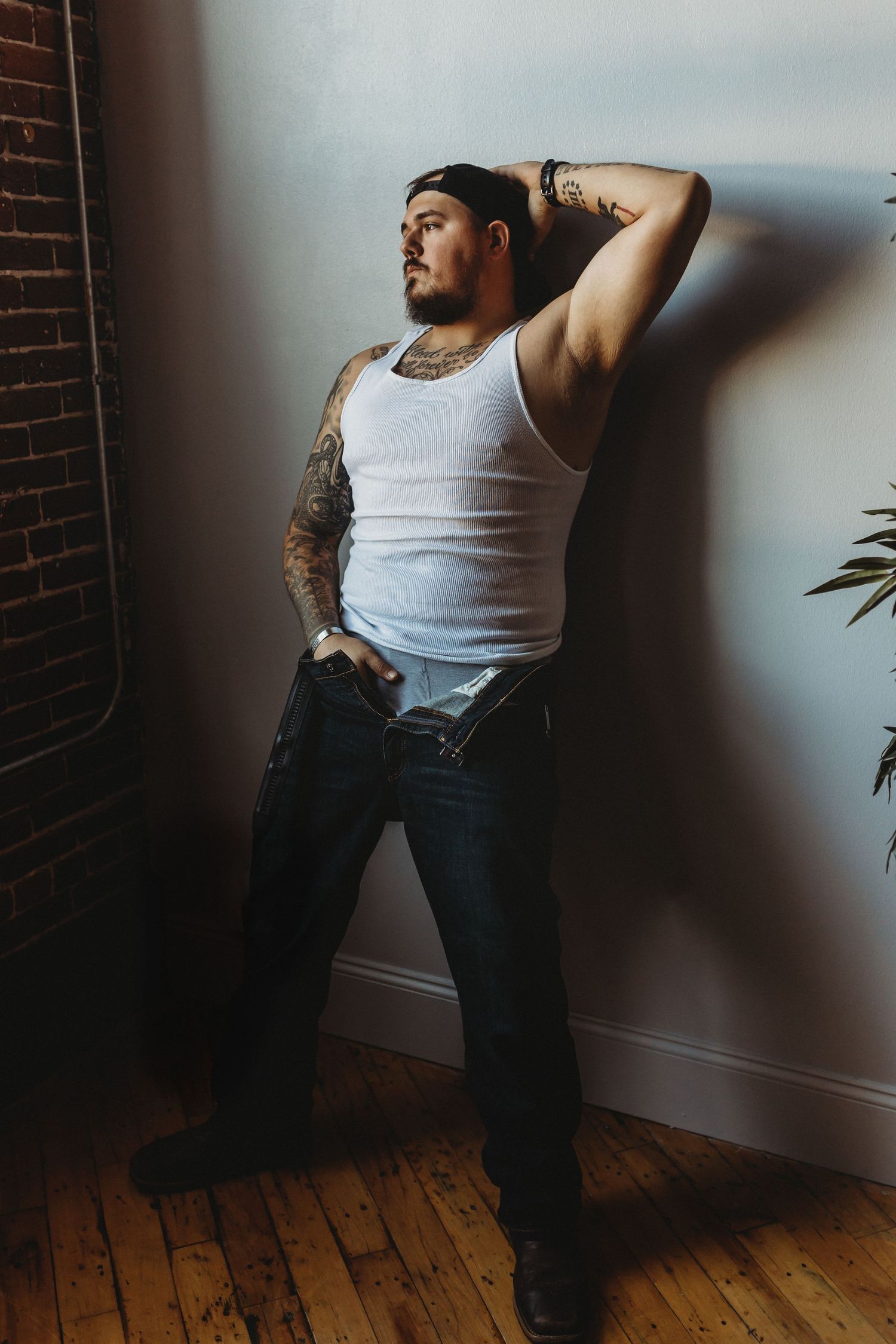 Athletic figure in white tank top and jeans poses against wall showing detailed arm tattoos.