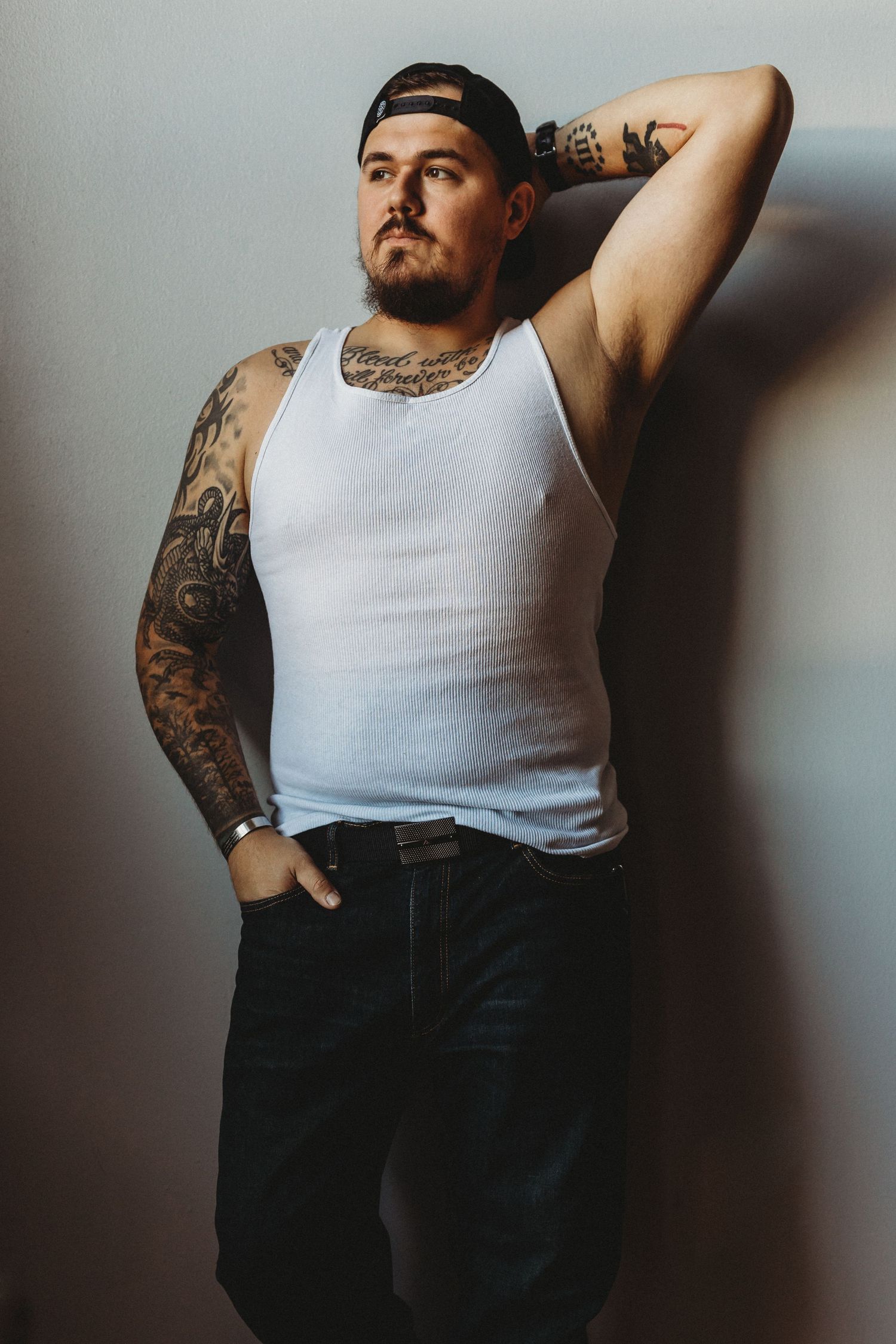 Studio portrait of person wearing white tank top and jeans posing with hand in pocket and tattooed arms.