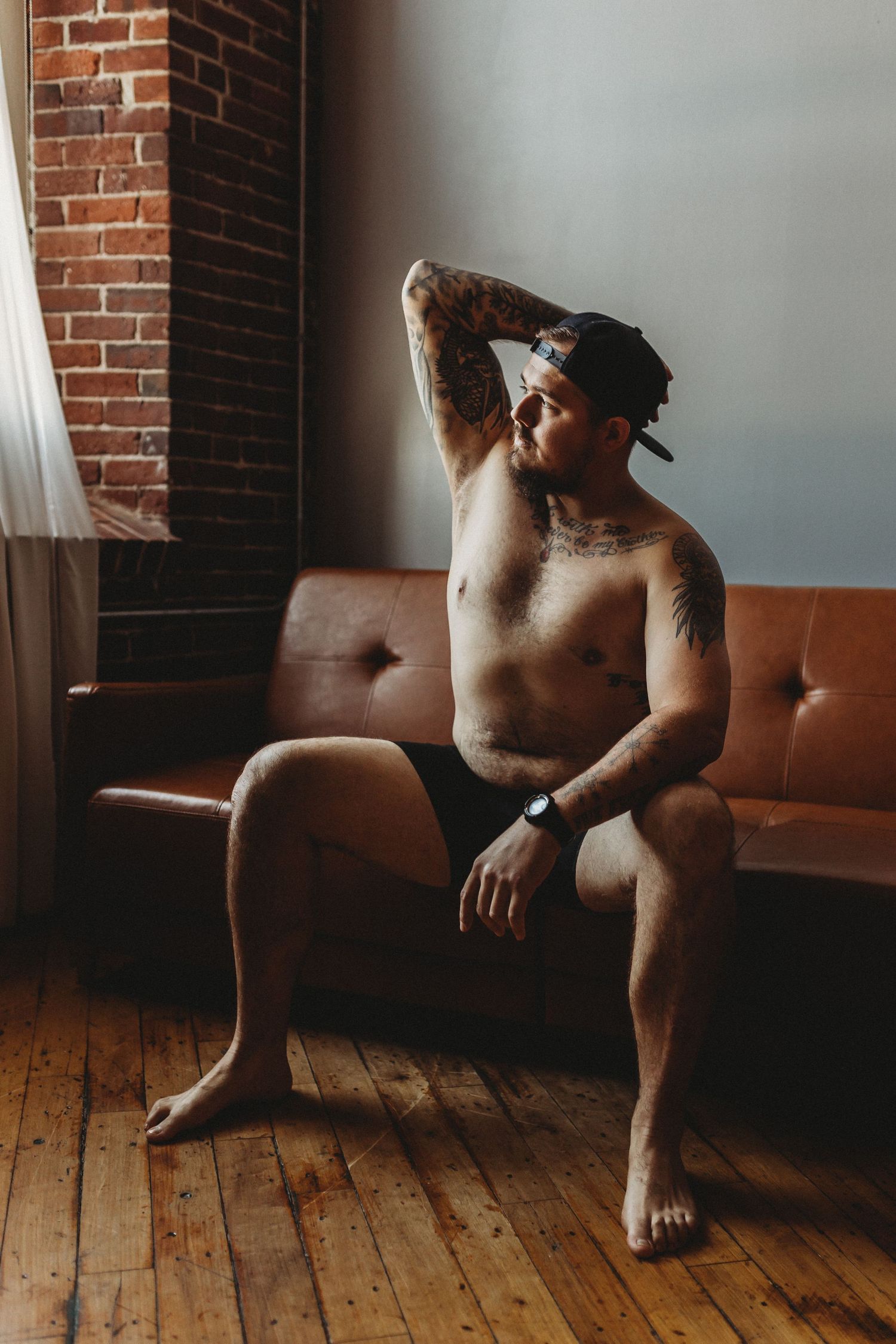 Artistic boudoir photo showing tattooed figure seated on brown leather couch.