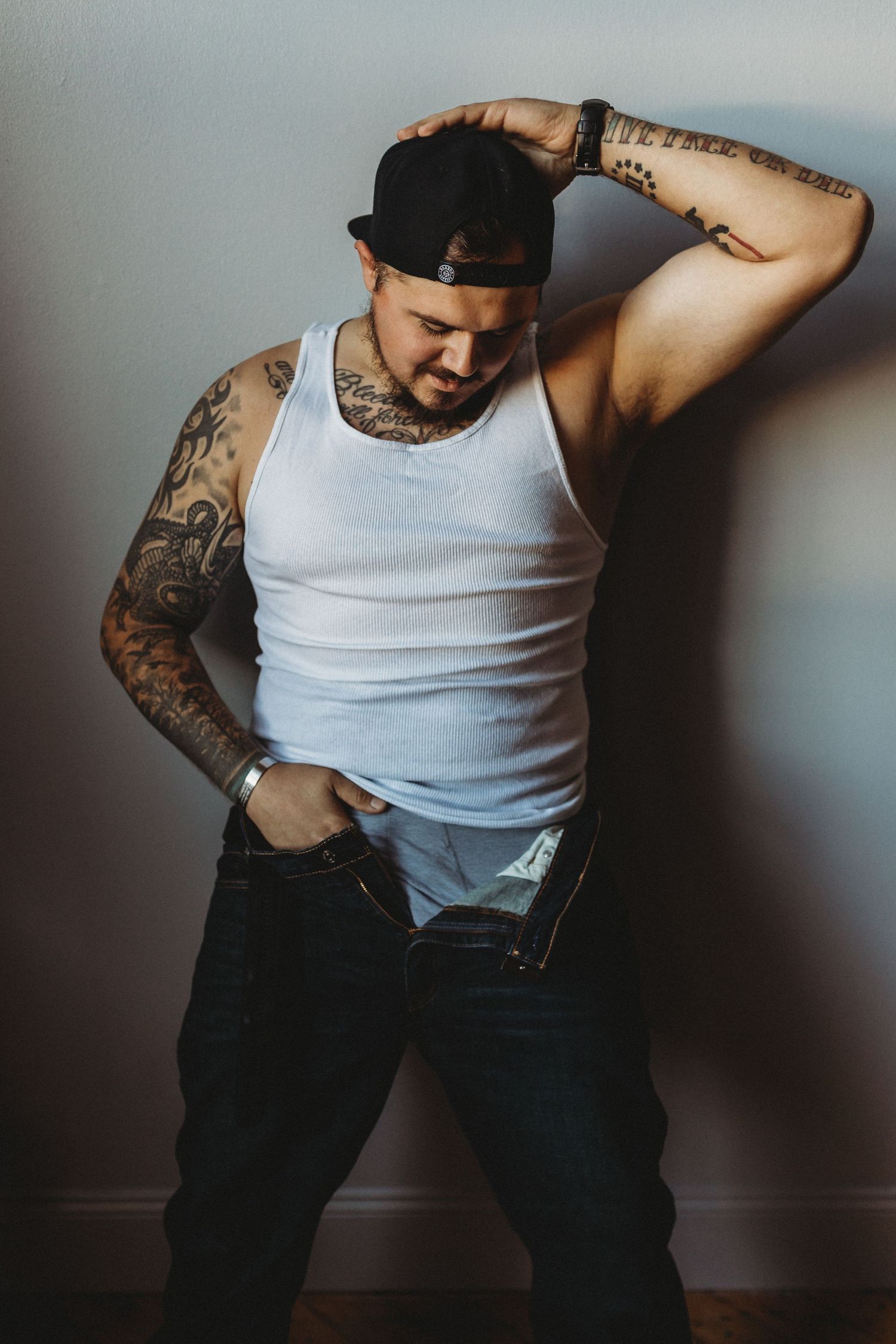 Tattooed person in backwards cap and tank top poses dramatically in moody lighting.